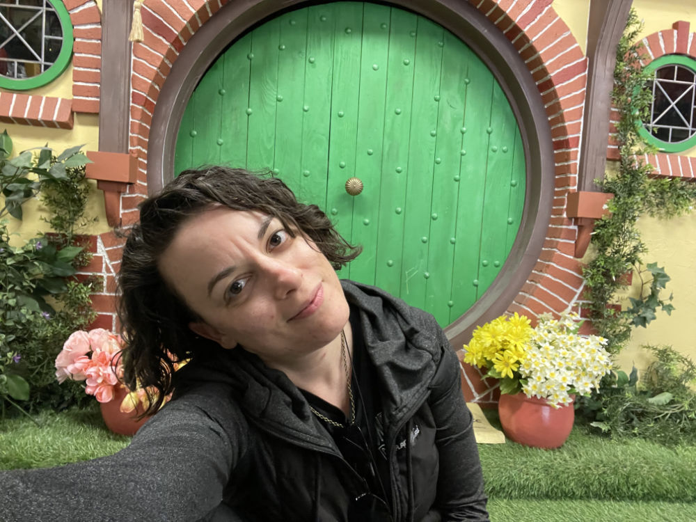 Bex, a white, gender non-conforming woman, sits and slightly grins, head slightly tilted. Bex is sitting in front of a Hobbit hole set with a large, green, round door, an arch of brick, and various flowers that look like a small garden or window boxes. Bex has mid length, dark hair and wears a gray jacket. Her face and posture suggest calm contentment.