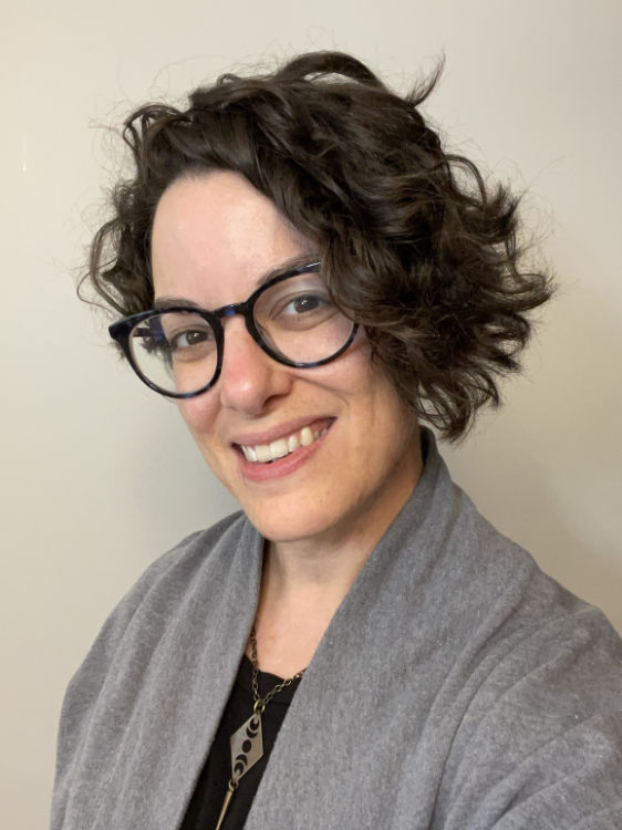 Bex, a white, gender non-conforming woman, grins toward the camera in front of a beige backdrop. Bex has mid length, dark hair and wears glasses and a gray cardigan over a black shirt. Her face and posture suggest joyful confidence and openness.