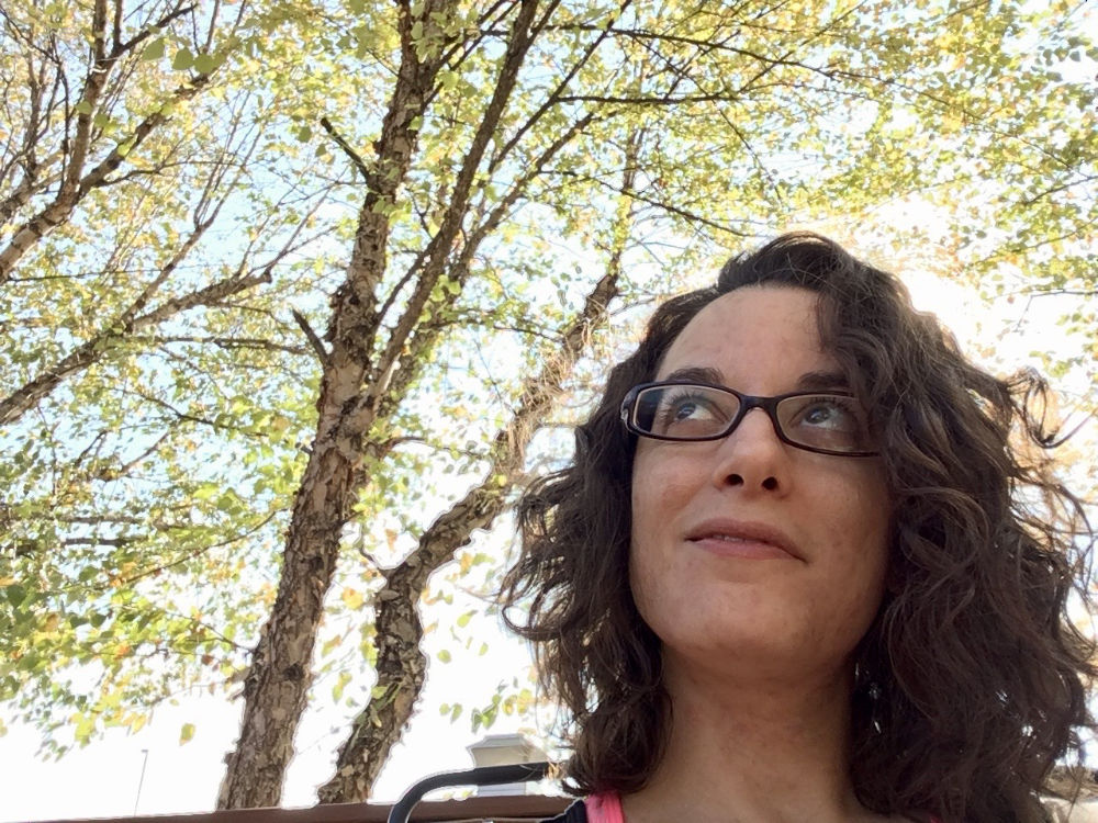 Bex, a white, gender non-conforming woman, grins upwardly toward the sky. Bex's face and neck are visible in frame; she has mid length, dark, curly hair and wears glasses. Behind her is a vibrantly green tree with branches, sky, and sunlight filling the rest of the frame. Her face and posture suggest joyful optimism.